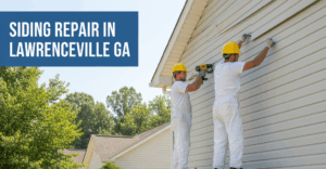 Two construction workers in yellow hard hats and white overalls are on scaffolding, repairing white vinyl siding on a house under a clear sky. A blue tarp on the grass below is covered with siding pieces and tools.